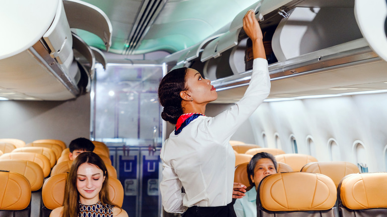 A flight attendant secures an overhead bin in a modern airplane cabin as passengers settle into their seats