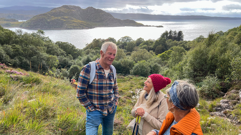 Three hikers smile and chat in a grassy clearing above a lake on a cloudy day in Scotland