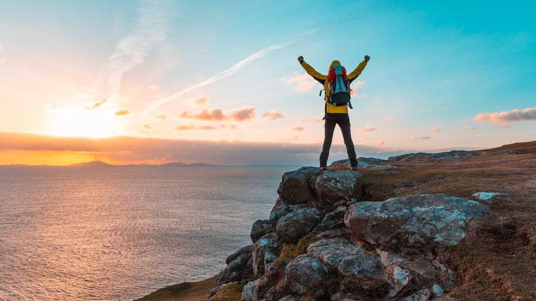 A hiker raises their arms triumphantly on a rocky ledge overlooking a lake and sunset in Scotland