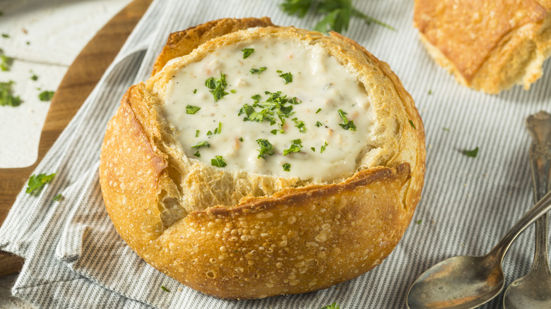 Clam Chowder in a sourdough bread bowl topped with parsley sitting on top of a striped towel and next to two spoons.