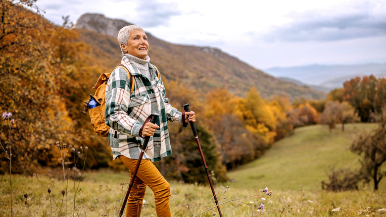 Senior woman with short grey hair and plaid shirt hiking in the mountains with yellow backpack and walking sticks