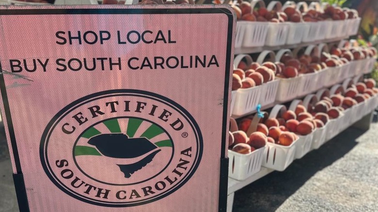 A sign that reads "shop local buy South Carolina" with baskets of ripe peaches behind it