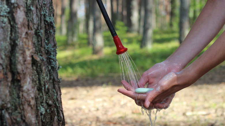 A person cups water pouring from a camping shower and a bar of soap in the woods.