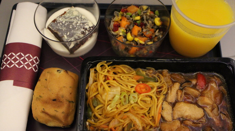 A tray of airplane food including stir-fry noodles, orange juice, salad and desert with a roll of bread and utensils wrapped in a napkin