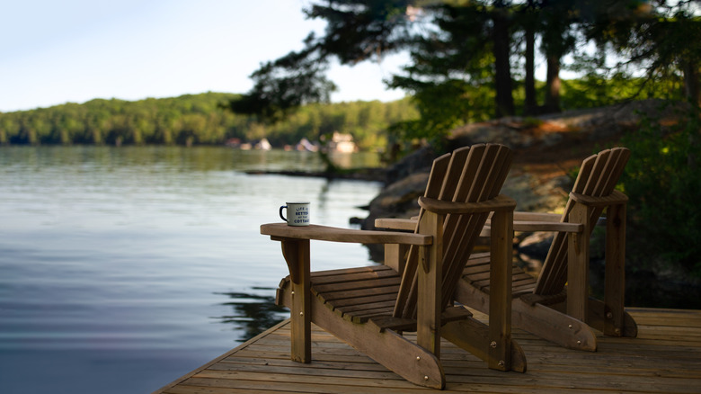Adirondack chairs face a lake on a calm morning with trees in the background.