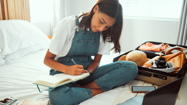 A young woman writing notes with her luggage and laptop in bed