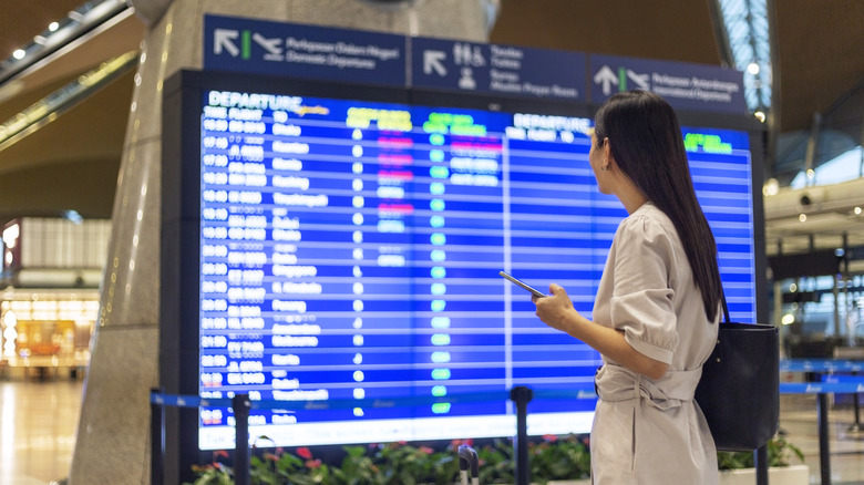 Woman with bag and phone looking at a departure board at the airport