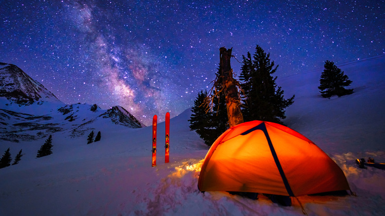 Night view of a ski mountaineer's camp, with a glowing tent and a pair of skis visible in the foreground, and the Milky Way galaxy visible in the background.