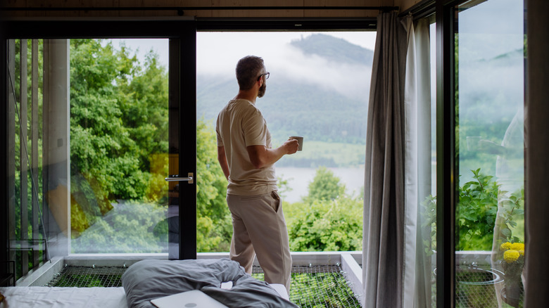 Man stands in hotel room overlooking mountain, holding coffee cup, with laptop on nearby unmade bed