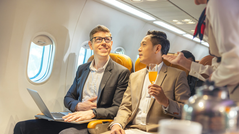 Two young men in suits sitting in an airplane getting served by a flight attendant