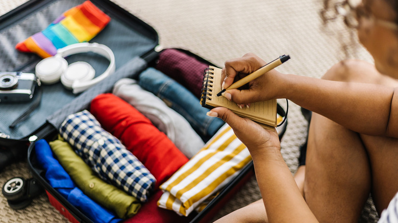 A woman writing a list while looking at luggage with colorful rolled-up clothes.