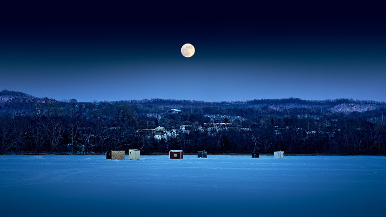 A full moon over a snowy mountain and a frozen lake with small, potable buildings on top