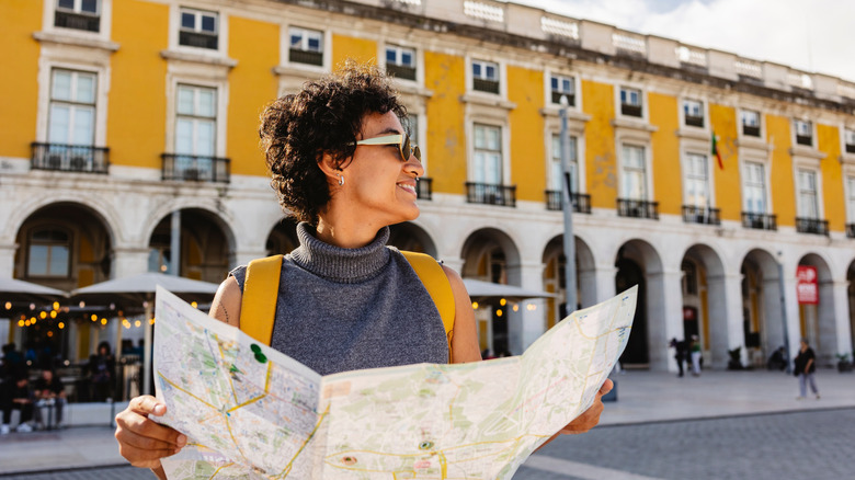 Smiling tourist is holding a map and wearing a yellow backpack while visiting the beautiful city of Lisbon in Portugal during summer vacations