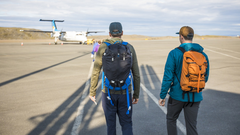 Two backpackers on a tarmac walking toward a small plane.