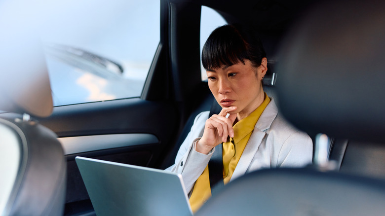 An Asian businesswoman in workclothes at the back of a car holding a laptop