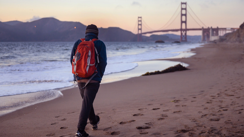 man with red backpack walking along a beach with the golden gate bridge in the background