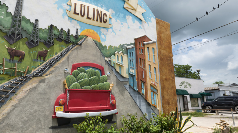 Colorful mural of a red truck carrying watermelons past railroad tracks, cattle and oil pumps in Luling