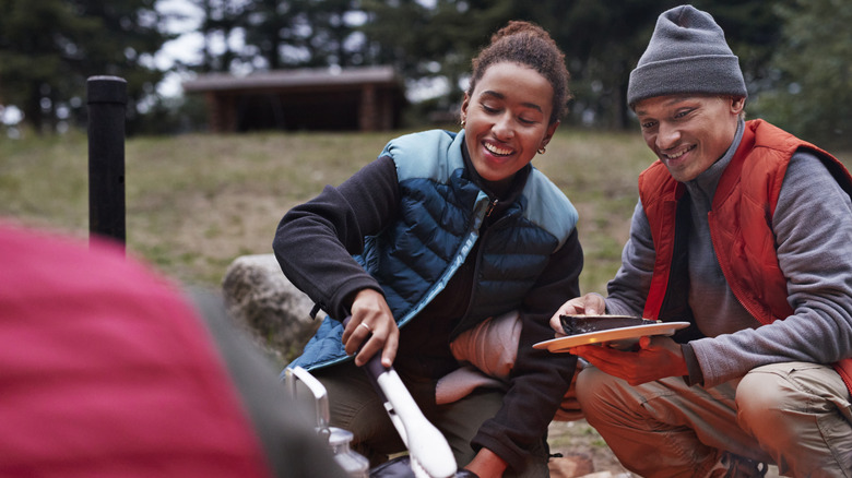 Smiling woman preparing food over a camp fire near a male friend.