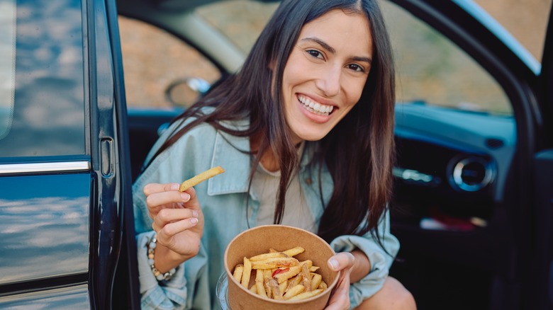 Woman in a car with the door open, smiling and eating French fries