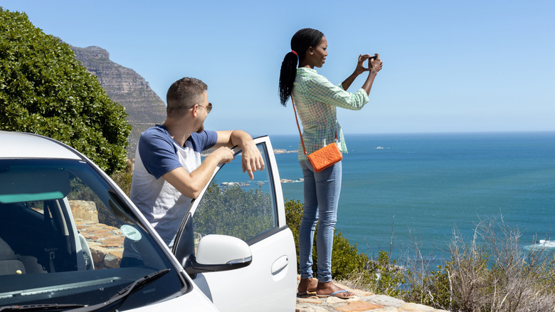 Couple taking picture outside car of ocean on road trip