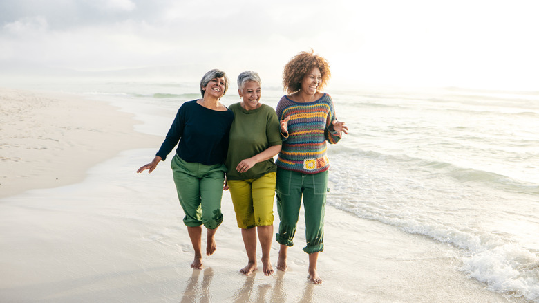 Three older women are smiling and laughing on a beach with the water from the ocean behind them