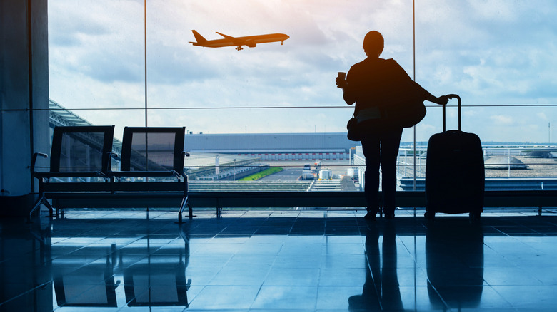 A woman is silhouetted by a window in an airport terminal, with a plane flying by in the background