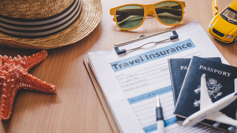 A clipboard with papers that read "travel insurance" with pen, passport, sunglasses and hat on top.