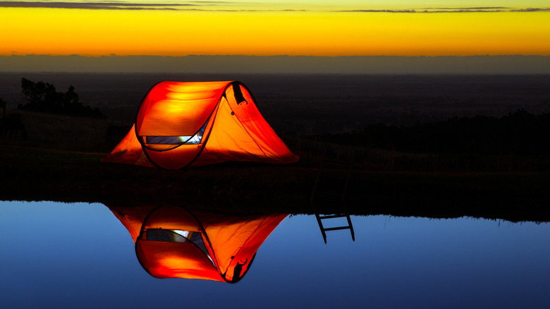 An orange tent reflected in a blue lake near sunrise with a yellow sky in the background.