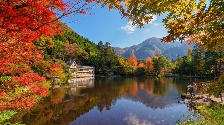 A river surrounded by a red, green, and orange autumnal landscape, with a house on one side and people on a dock.