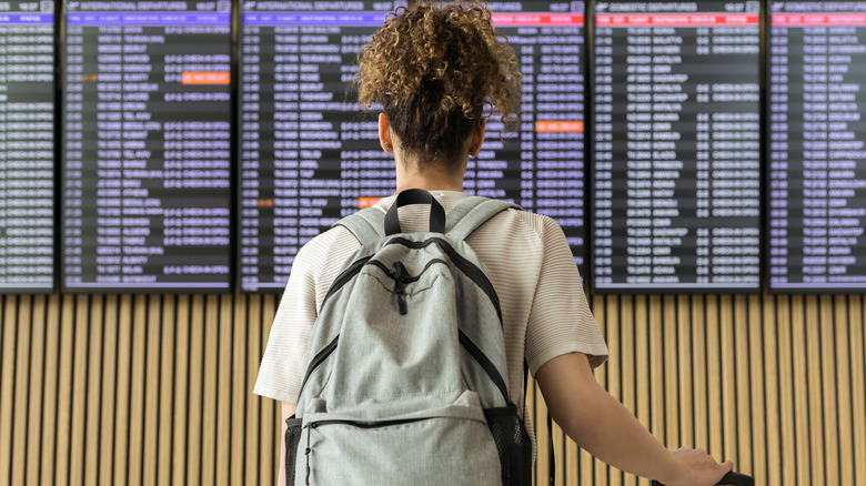 Backview of a woman with a backpack and luggage facing an airport departures and arrivals panel