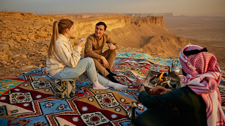 Tourist couple drinking traditional tea with a bedouin. Orange desert cliffs are in the background.