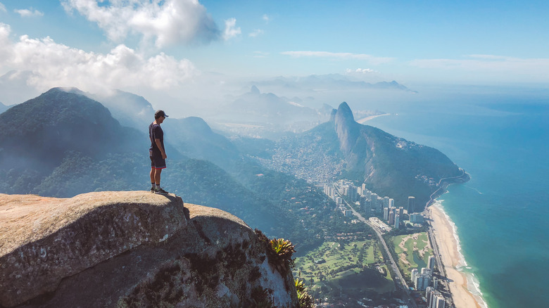 A man stands at the top of a seaside cliff, looking down onto a beach in Rio