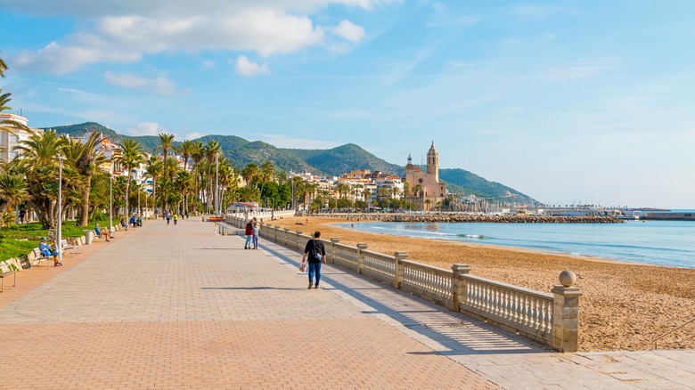 A beachside promenade extends towards a town with a church and mountains behind it