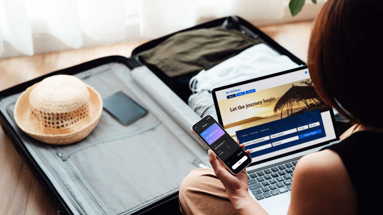 A woman has opened a digital credit card on her phone while booking a flight on her computer with her passport lying on a suitcase at her feet