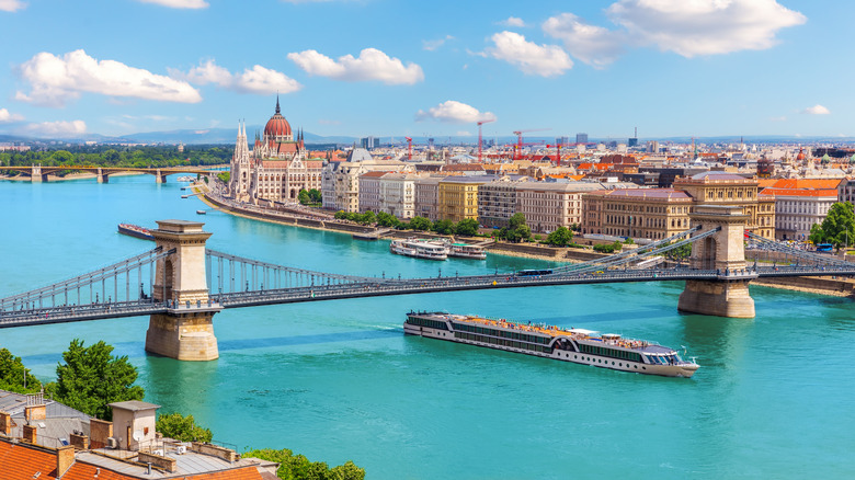 Aerial view of a boat cruising through the blue waters of the Danube River during the day, with Budapest's skyline and the Chain Bridge visible in the background