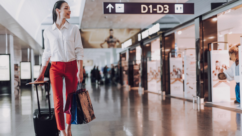 Stylish woman with suitcase walking past airport shops