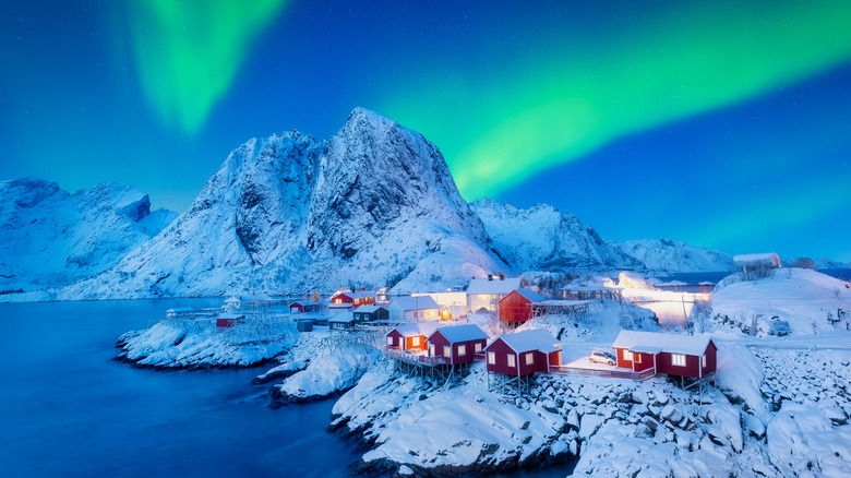 Winter shot of Hamnoy village in Norway with snow-covered mountains and the Northern Lights in the sky.