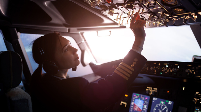Female pilot flying inside the cockpit adusting settings on the upper panel a sun shines outside