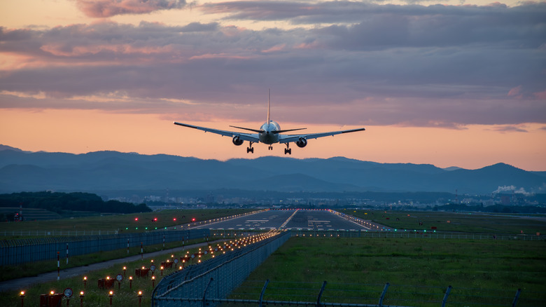 A jet plane lands at sunset on a lit runway in a grassy field with mountains in the background