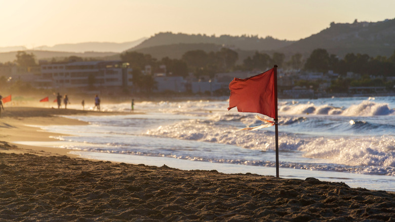Unrecognizable people walking on a beach with a red flag warning of big waves, sunny dawn