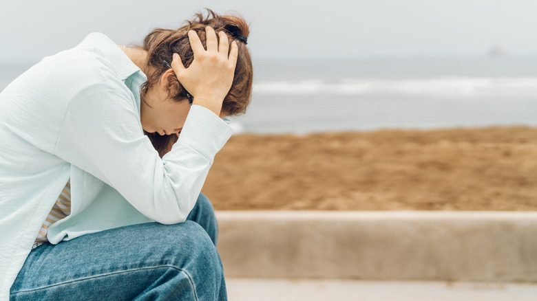 Worried young woman holding her head in front of the beach, sitting on a bench.