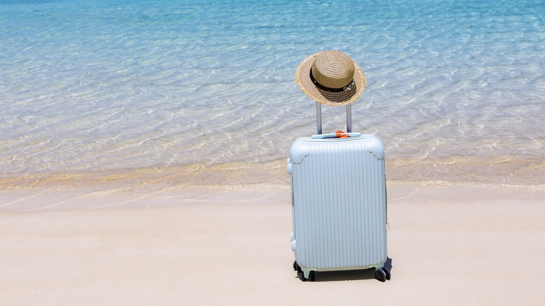 A light blue suitcase with wheels and a straw hat on a beach