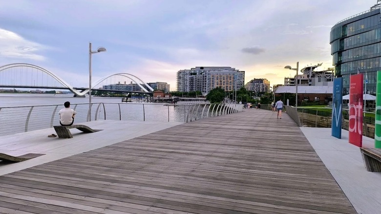 The Navy Yard Boardwalk Trail along the Anacostia River in Washington, D.C., with views of an arched bridge across the water and the skyline in the distance.