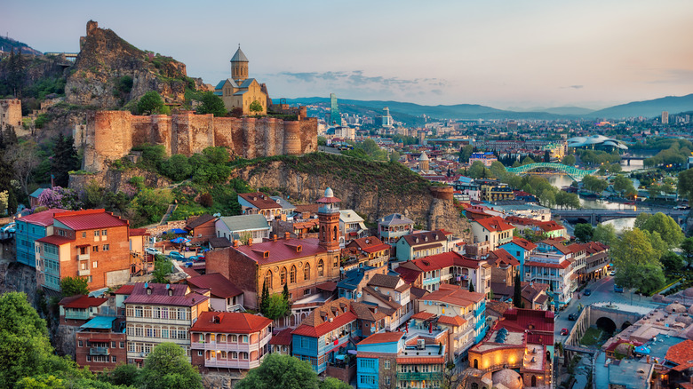 A town of red-roofed buildings built along the sloping terrain of a mountain, with a castle and fortifactions at the top of the mountain. More buildings feature in the background.