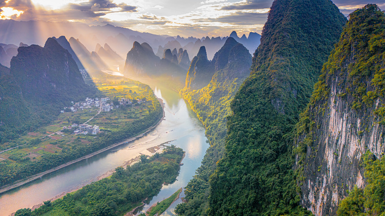 A river valley with mist-shrouded karst mountains on both sides of the water, lit overhead by a dramatic sunset. Houses dot one side of the river.