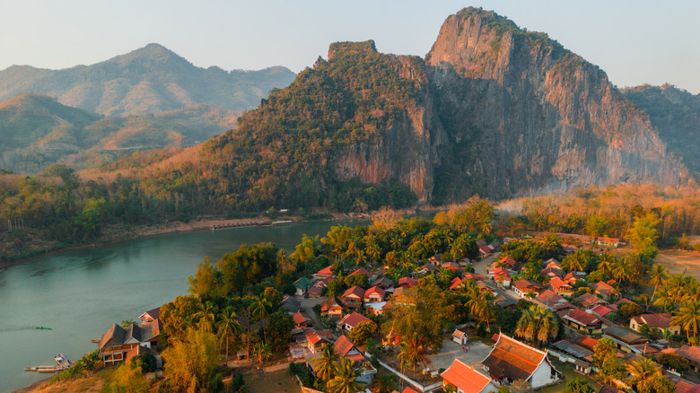 A village with concave, sloped red roofed houses surrounded by jungle. It is separated from mountains by a winding river.