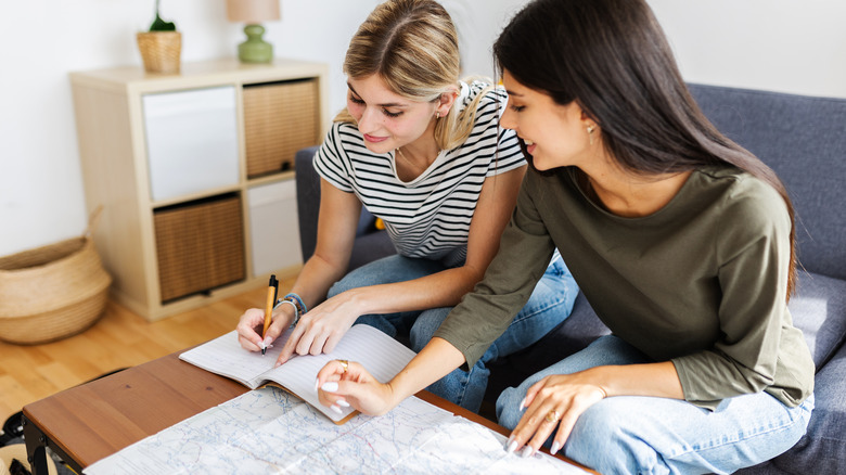 Two women sitting on a living room couch. One of them is writing on a notebook and the other one has a map in front of her.