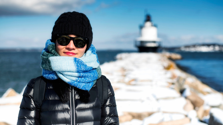 A woman in sunglasses and scarf stands in front of a New England jetty and lighthouse