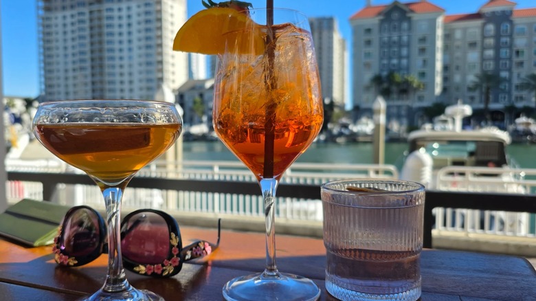 Cocktails and sunglasses on a table in Anchor & Brine's shaded patio looking out toward docked boats and the Tampa skyline across the channel