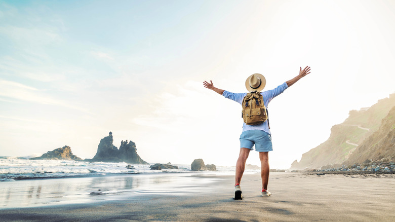 a man with a backpack standing with his arms thrown up in joy on the beach